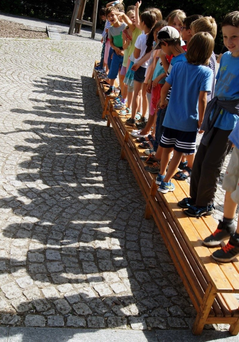 Kinder der ersten Klasse sitzen in einer Reihe auf der Bank vor dem Schulhaus
